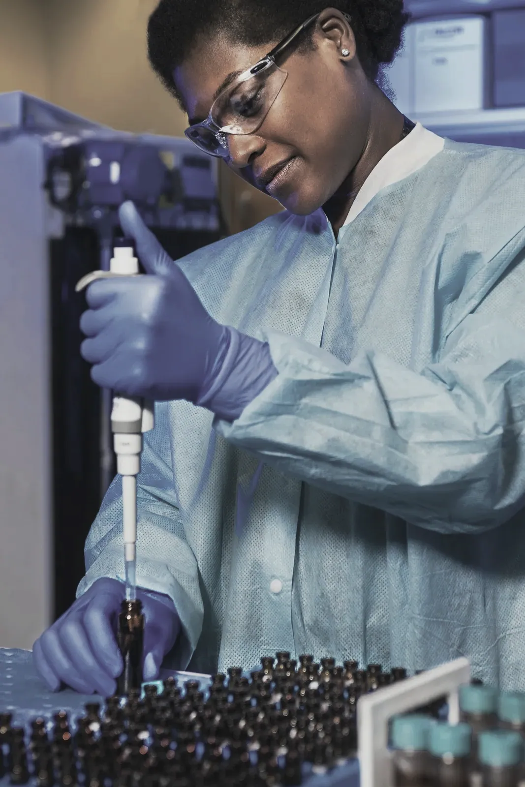 A laboratory technician in a blue gown and gloves using a pipette over a rack of sample vials