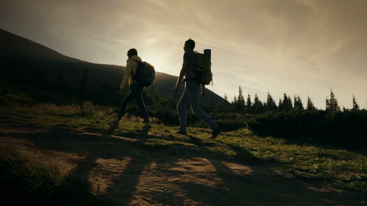 A man and a woman with packs hiking up a mountain trail into a low golden sun, moving with easy unhurried strides, the left of the frame falling into shadow.