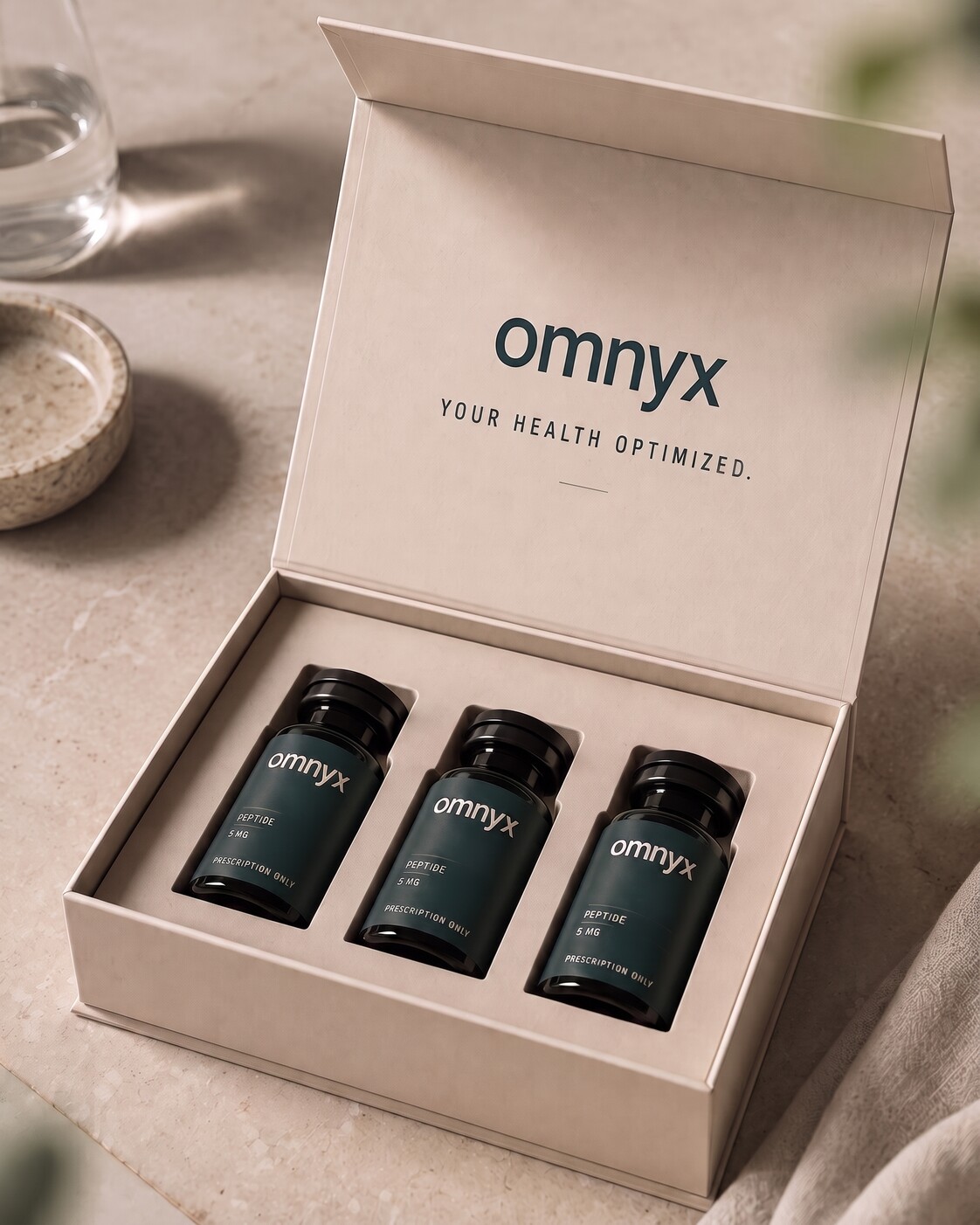 An open omnyx kit on a stone surface in soft daylight, three branded bottles seated inside the bone-colored box, suggesting concierge care delivered to the home.