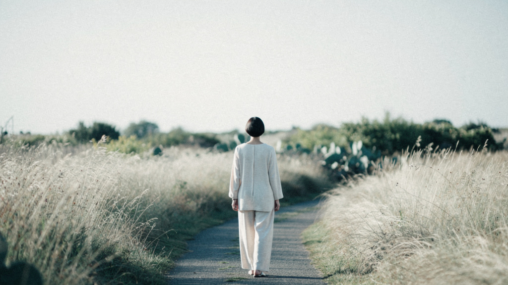 A person walking at ease through tall pale grass in soft morning light, seen from a distance.