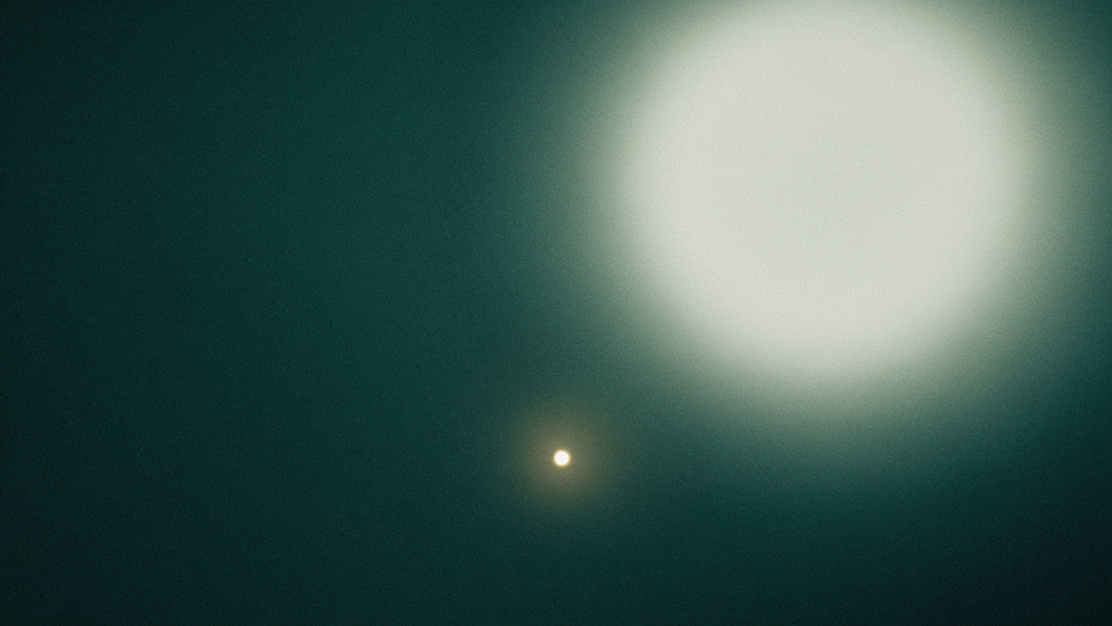 One small, low light holding steady in a field of teal darkness, quiet and sufficient, beside the washed-out glare of a far brighter one that adds nothing.