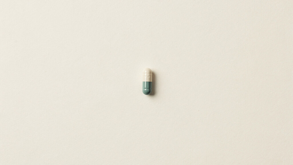 A solitary pale capsule resting at the center of a wide, quiet bone colored expanse, small against the surrounding emptiness, rendered in soft matte tones with a single muted teal accent set slightly apart.