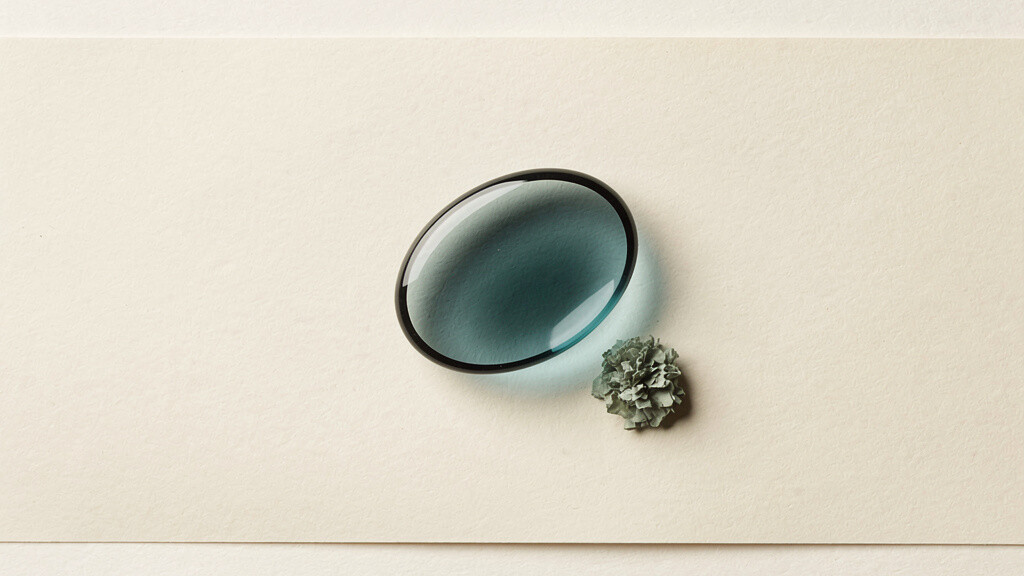 A single outsized translucent teal capsule shape hovers over a warm bone field, nearly hollow inside, while a small dense cluster of matcha sage rests beneath it, the loud promise of the pill above the small real signal of food.